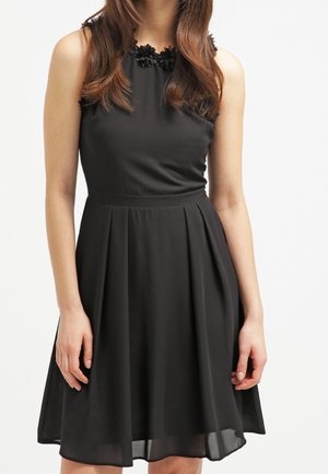 Nainen, joka käyttää hihattoman, polvipituisen mustan mekon, jossa on kukkakuvioita kaula-aukossa, seisoo käsivarret rentoina sivuillaan.