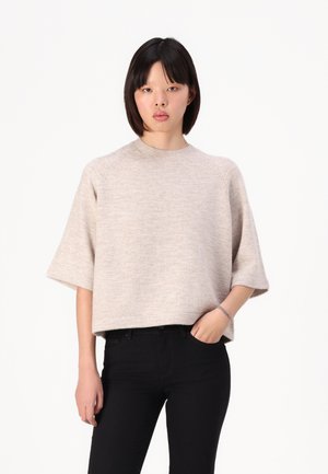 Beige, kortermet genser med ribbestrikket hals og løs passform, båret over svarte jeans, med en myk tekstur.