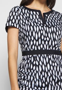 Robe noire à manches courtes ornée d'un motif de feuilles blanches, avec un col en V subtil et une ceinture noire contrastante. Texture de tissu lisse.