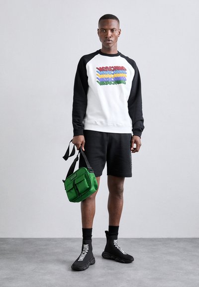 Czarna i biała bluza z kolorowym logo, czarne szorty, zielona torba, czarne skarpetki z napisem i czarne buty z falistą podeszwą.