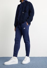 Navy corduroy jacket, navy hoodie, matching joggers with white Nike logo, and white sneakers with textured soles on a blue carpet background.