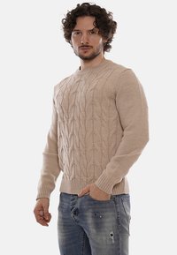 Uomo con capelli ricci che indossa un maglione beige con trecce e jeans blu strappati, in piedi con una mano in tasca.