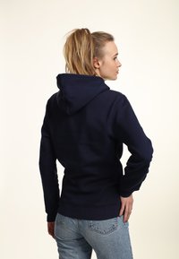 Felpa blu navy realizzata in tessuto morbido, dotata di un ampio cappuccio, maniche lunghe e polsini e orlo a coste. Indossata con jeans blu chiaro.