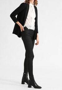 Blazer negro con mangas arremangadas sobre una blusa de encaje blanca, combinado con jeans ajustados negros y botines de tacón. Textura suave y diseño entallado.