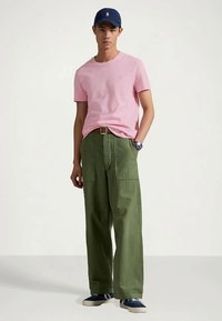 Νεαρός άνδρας που φοράει ροζ μπλουζάκι, ελαιοπράσινα φαρδιά παντελόνια, ναυτικό καπέλο, μπλε αθλητικά παπούτσια και ρολόι, έχοντας φόντο απλή επιφάνεια.