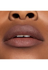 Rouge à lèvres mat dans une teinte taupe chaude, mettant en valeur des lèvres pulpeuses avec une surface texturée et une légère brillance sur la lèvre inférieure, sur un fond de peau lisse.