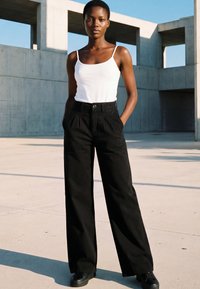 Jeune femme debout en extérieur, portant un pantalon large noir, un haut blanc sans manches et des chaussures noires, sur fond d'architecture moderne en béton.