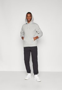 Gray hoodie with a front pocket and drawstring hood, paired with black tapered pants and white sneakers, worn by a person standing upright.