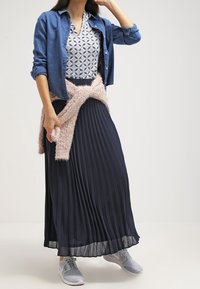 Jeansskjorta, mönstrad topp, plisserad navy maxikjol, fluffig blush-wrap och grå sneakers. Texturerna kontrasterar; färgerna är mjuka och dämpade.