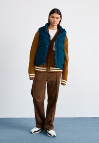 Teal pufferväst över en brun cardigan med vita ränder, bruna byxor och vita sneakers med gröna accenter. Släta texturer, avslappnad passform.