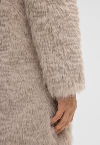 Mano appoggiata sul lato di un indumento a maniche lunghe beige e fuzzy, che mostra i dettagli del tessuto e il contrasto del tono della pelle su uno sfondo chiaro.