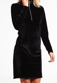 Schwarzes Samt-Kleid mit langen Ärmeln, Hochkragen und halbem Reißverschluss. Die glatte Textur und die taillierte Silhouette betonen das Design.