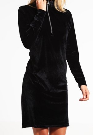Svart sammet långärmad klänning med hög hals och halvdragkedja. Den släta texturen och figurnära silhuetten framhäver designen.