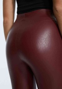 Leggings in finta pelle color bordeaux con una texture liscia, design aderente, cuciture visibili e vita alta.