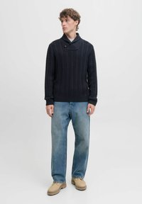 Granatowy sweter z dzianiny z grubym wzorem w warkocze, z kołnierzem typu szal i ściągaczami, w połączeniu z jasnoniebieskimi dżinsami i beżowymi butami.