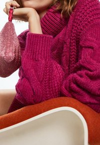 Une femme portant un pull épais magenta tient un petit sac à main rose orné de perles, assise sur une chaise moderne orange et blanche.