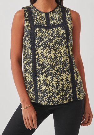 Bezrekawnik w żółtym kolorze z czarnym kwiatowym wzorem i pionowymi czarnymi koronkowymi panelami, noszony z czarnymi spodniami i złotą bransoletką na lewej ręce.