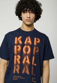 T-shirt en coton bleu marine avec un grand texte orange en spray "KAPORAL" et un texte supplémentaire sur le bas à l'avant. Manches courtes, col rond.