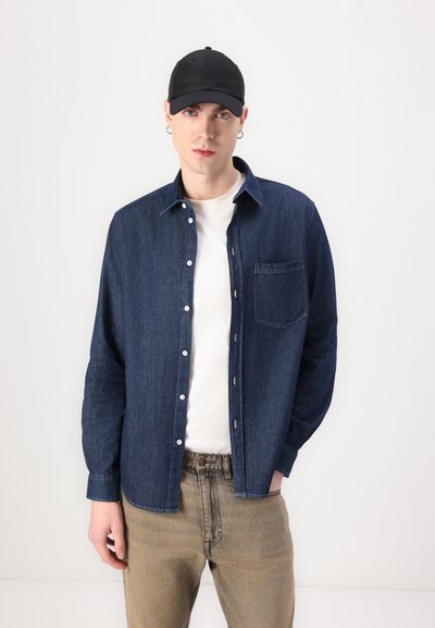 Chemise en denim bleu foncé à boutons avec une poche poitrine, associée à un T-shirt blanc et un pantalon beige, le tout complété par une casquette noire.