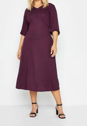 Mujer con vestido midi de color púrpura oscuro, con mangas de tres cuartos y sandalias negras de tacón, sobre un fondo claro y liso.