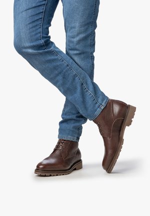 Bottes de cheville en cuir marron avec une finition texturée, bout rond et semelle en caoutchouc marron. Associées à un jean en denim bleu, les jambes croisées au niveau de la cheville.
