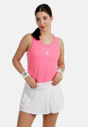 Jeune femme ajustant une jupe de tennis blanche, portant un haut sans manches rose et des bracelets blancs, posant devant un fond uni.