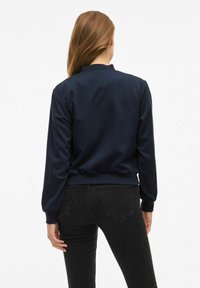 Veste de pilote marine en tissu lisse avec des poignets et un ourlet côtelés. Présente un col montant et un design simple et structuré.