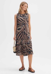 Maxi dress with a navy base, featuring a brown and white abstract pattern, sleeveless design, and tiered hem. Worn with sandals and a woven bag.