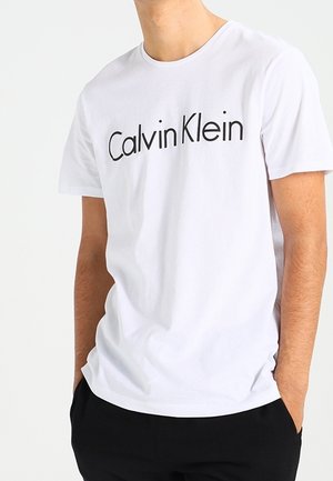 Mężczyzna ubrany w białą koszulkę z krótkim rękawem z czarnym logo "Calvin Klein", ręce w kieszeniach, stojący na jasnym, jednolitym tle.