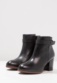 Botas de cuero negras hasta el tobillo con un acabado texturizado, tacón bloque de altura media y correas anchas alrededor del tobillo. Detalle de cremallera en el lado.