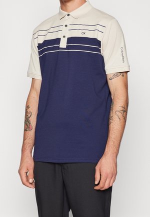 Mężczyzna ubrany w koszulkę polo z krótkim rękawem, z beżową górą, granatowym dołem, poziomymi białymi paskami oraz logo Calvin Klein na piersi i rękawie.