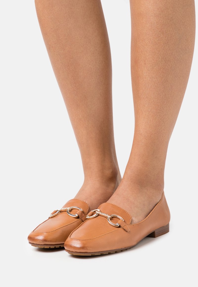 Mocassins en cuir marron clair avec un détail argenté en forme de mors de cheval, portés pieds nus sur un fond neutre.