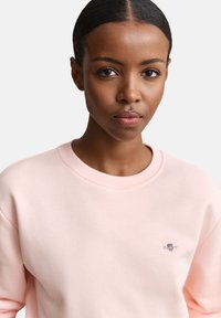 Γυναίκα με λείο, χτενισμένο προς τα πίσω μαλλί φοράει ένα ανοιχτό ροζ φούτερ με μικρό κεντημένο λογότυπο στο στήθος, ουδέτερο φόντο.