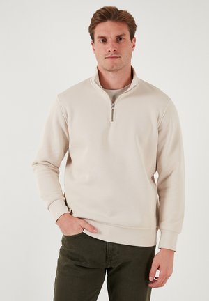 Jasno beżowy półzapinany sweatshirt z długimi rękawami i prążkowanymi mankietami, wykonany z miękkiego materiału, z stójką i gładką fakturą.
