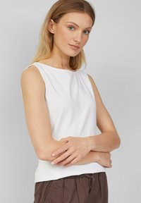 Top blanco sin mangas con escote redondo, textura suave y una ligera forma en A. Combinado con pantalones marrones con cordones.