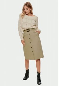Suéter de malha bege com padrão de pontos abertos, combinado com uma saia cáqui até ao joelho com cinto, botões e bolsos laterais. Botas pretas até ao tornozelo.