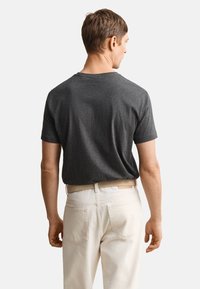 Mann trägt ein dunkelgraues kurzärmeliges T-Shirt, das in eine cremefarbene Hose mit beigem Gürtel gesteckt ist, und schaut mit dem Rücken zu einem einfarbigen Hintergrund.
