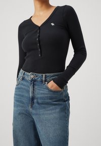Sort ribbet langærmet skjorte med V-hals og knapdetalje, med et lille hvidt logo på brystet, parret med lyseblå denimjeans.