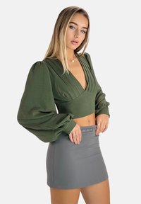Grünes langärmliges Crop-Top mit geschichteter Vorderseite und voluminösen Ärmeln, kombiniert mit einem figurbetonten grauen Minirock mit einem Gürteltail.