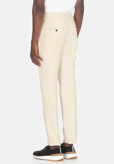 Pantaloni beige con una vestibilità affusolata, con tasche posteriori e fondo elastico. Indossati con sneakers nere. Tessuto dalla texture morbida.