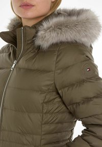 Giacca puffa oliva con cappuccio staccabile foderato in pelliccia, design a righe verticali, zip argentata e toppa rotonda con logo sulla manica.