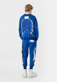 Conjunto de sudadera y pantalones azules que presenta gráficos de alas en blanco y azul en la parte posterior. El material es suave con una textura acogedora. Completado con botas blancas.