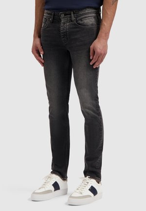 Man wearing slim-fit faded black jeans and white low-top sneakers with dark blue accents, standing against a plain light background.