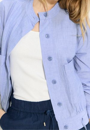 Vrouw draagt een lichtblauw jasje met knopen over een wit topje en donkerblauwe broek met trekkoord, met los haar over de schouder.