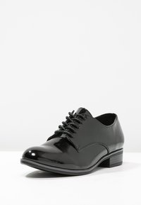 Chaussure de cérémonie en cuir verni noir avec un bout rond, des lacets à l'avant et un petit talon empilé. Texture lisse et détails de couture minimalistes.