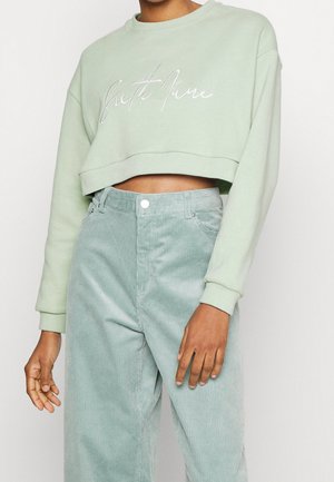 Personne portant un sweat court vert clair avec une inscription blanche et un pantalon en velours côtelé vert clair taille haute sur un fond uni.