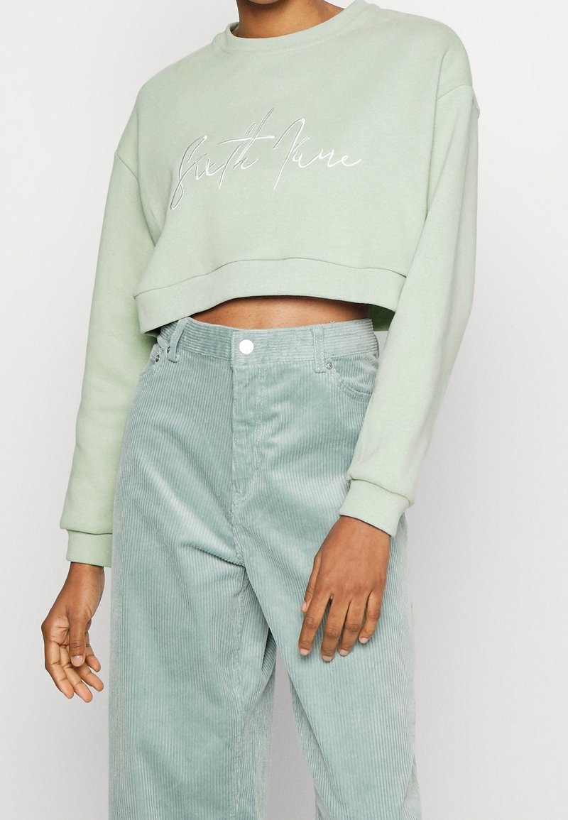 Personne portant un sweat court vert clair avec une inscription blanche et un pantalon en velours côtelé vert clair taille haute sur un fond uni.