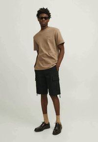 Giovane uomo con occhiali da sole, t-shirt beige, pantaloni cargo neri, calze beige e scarpe eleganti nere, in piedi con le mani nelle tasche.