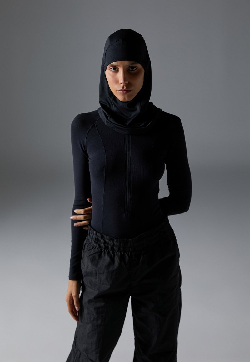 Svart atletisk langermet topp med høy krage og integrert hijab, kombinert med løse svarte bukser, på en nøytral bakgrunn.