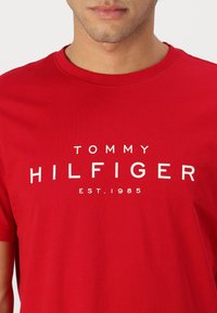 Raudona medvilninė marškinė su apvaliu kaklu. Balti žodžiai skelbia "TOMMY HILFIGER EST. 1985." Paprastas dizainas, standartinė išpjova, be matomų raštų ar akcentų.